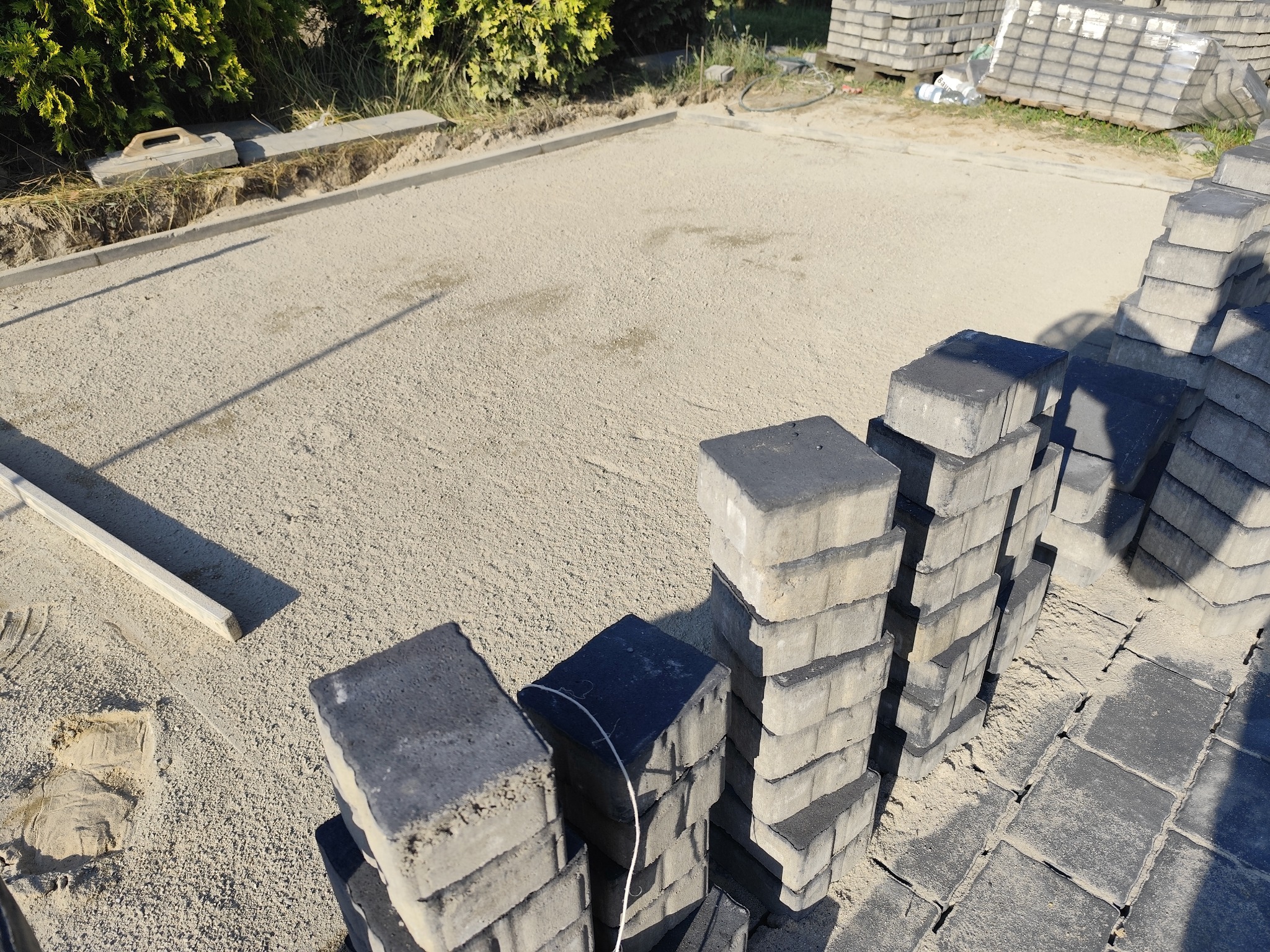Przygotowany teren pod kostkę brukową z ułożonymi obrzeżami, podsypką piaskową i stosem kostki gotowej do ułożenia. Widoczne narzędzia brukarskie.
