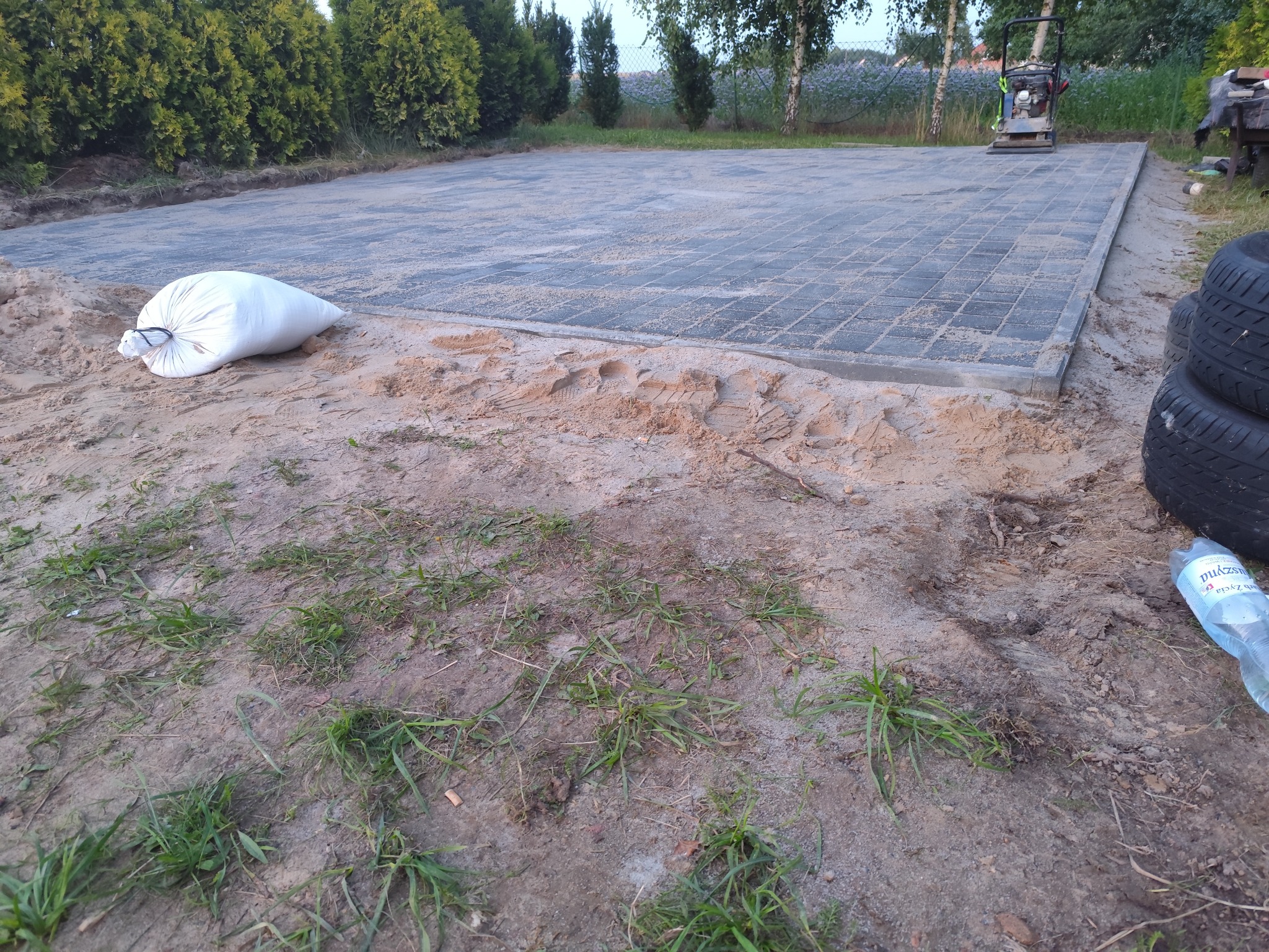 Połowa powierzchni wyłożona szarą kostką brukową, obok biały worek z piaskiem i ubijarka. Widoczne ślady stóp na piasku. W tle zieleń.