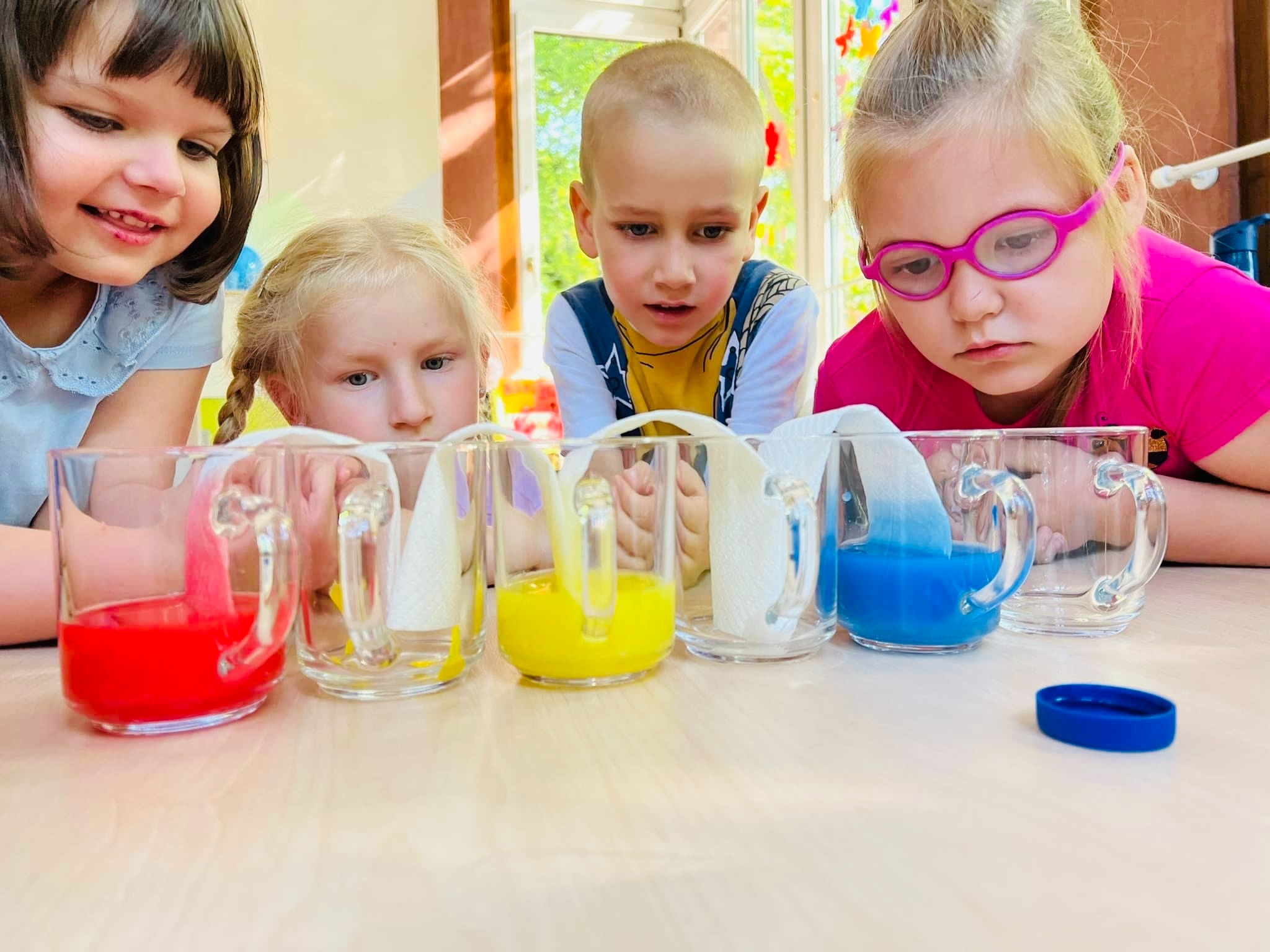 Czterech podekscytowanych przedszkolaków pochyla się nad stołem, obserwując szklane dzbanki z kolorową wodą: czerwony, żółty i niebieski, z ręcznikami papierowymi w środku.