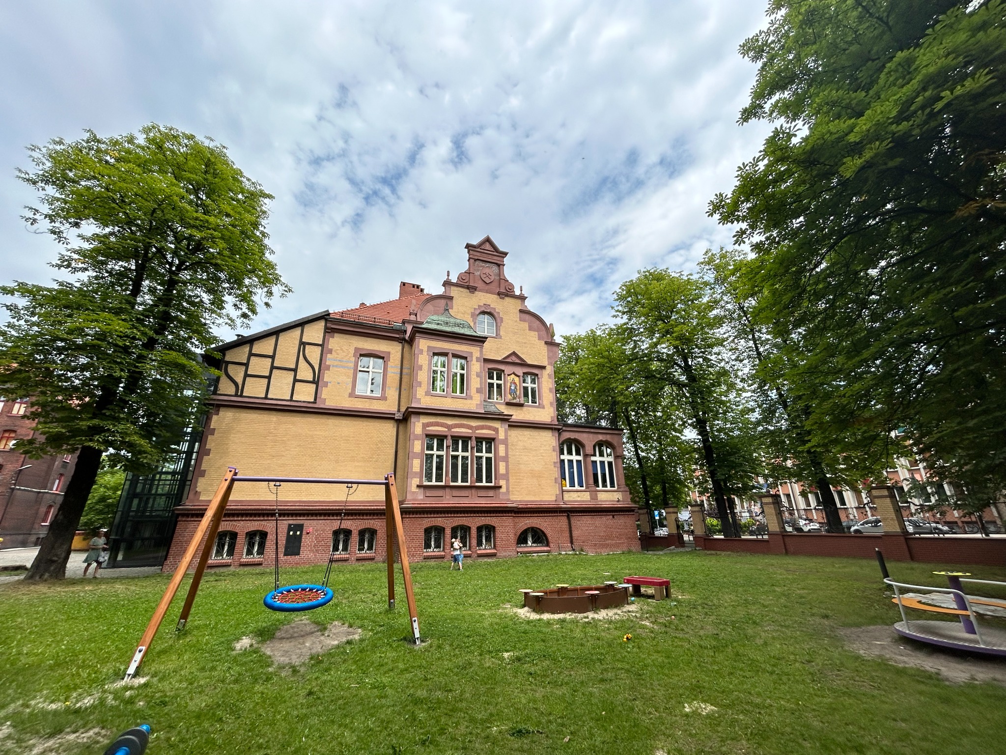Plac zabaw z huśtawką typu bocianie gniazdo i piaskownicą na tle budynku z żółtej cegły z czerwonymi elementami architektonicznymi, otoczony zielenią.