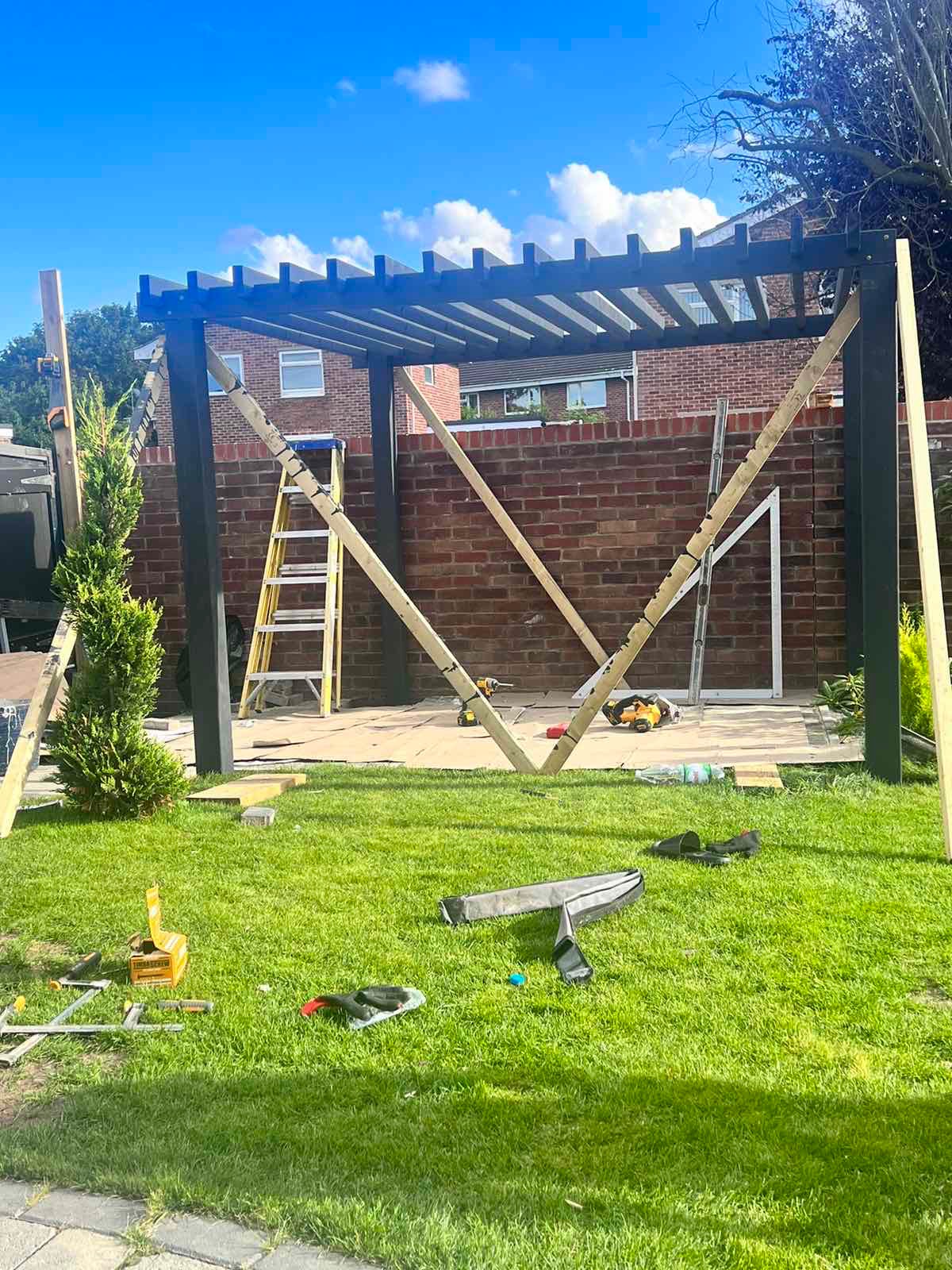 Montaż czarnej pergoli w ogrodzie z użyciem drewnianych podpór i drabiny, na trawie leżą narzędzia i akcesoria.