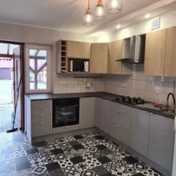 Nowoczesna kuchnia z szarymi szafkami, drewnianymi górnymi szafkami, czarnym blatem i wzorzystą czarno-białą podłogą. Nad blatem kuchennym białe kafelki. Widoczne trzy lampy wiszące w stylu...