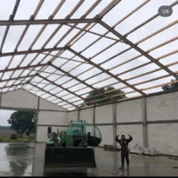 Konstrukcja dachu hali w trakcie budowy, widoczna więźba dachowa z belek, bloczki betonowe ścian i pracownik w uprzęży oraz zielona ładowarka teleskopowa.