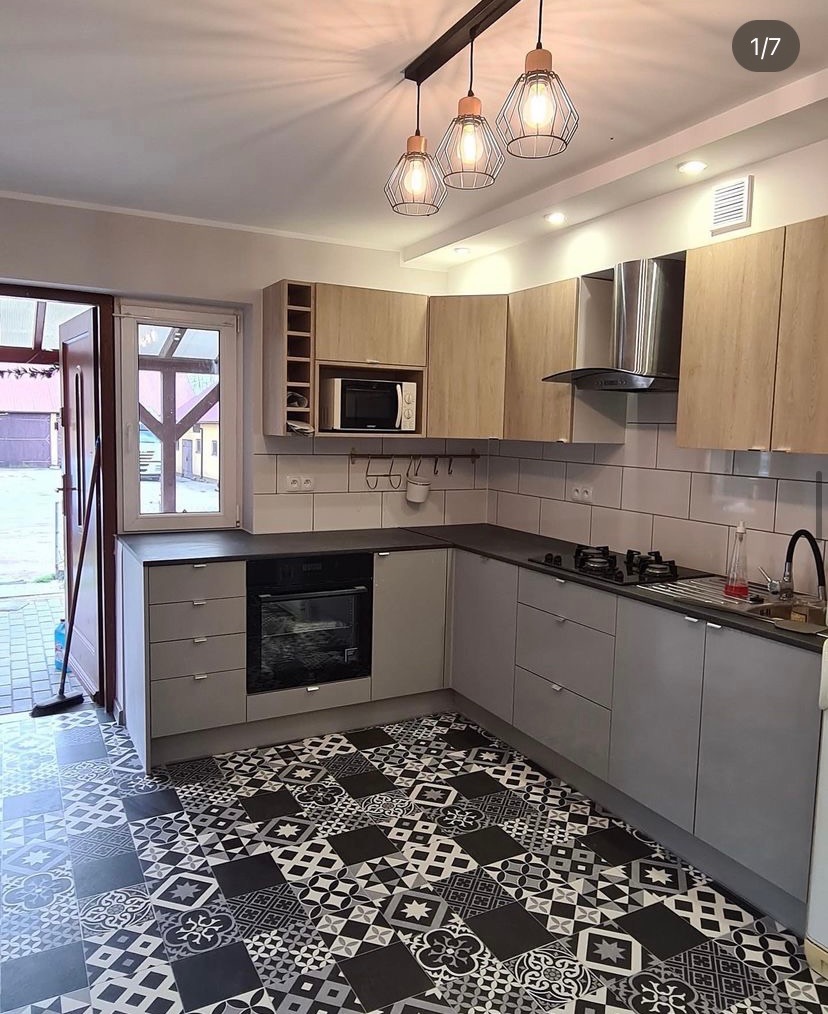 Nowoczesna kuchnia z szarymi szafkami, drewnianymi górnymi szafkami, czarnym blatem i wzorzystą czarno-białą podłogą. Nad blatem kuchennym białe kafelki. Widoczne trzy lampy wiszące w stylu...