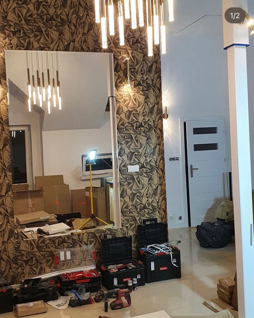 Wnętrze w trakcie prac wykończeniowych z widocznym lustrem, narzędziami, kartonami i lampą podłogową, ściana z dekoracyjną tapetą, sufit z nowoczesnym oświetleniem.