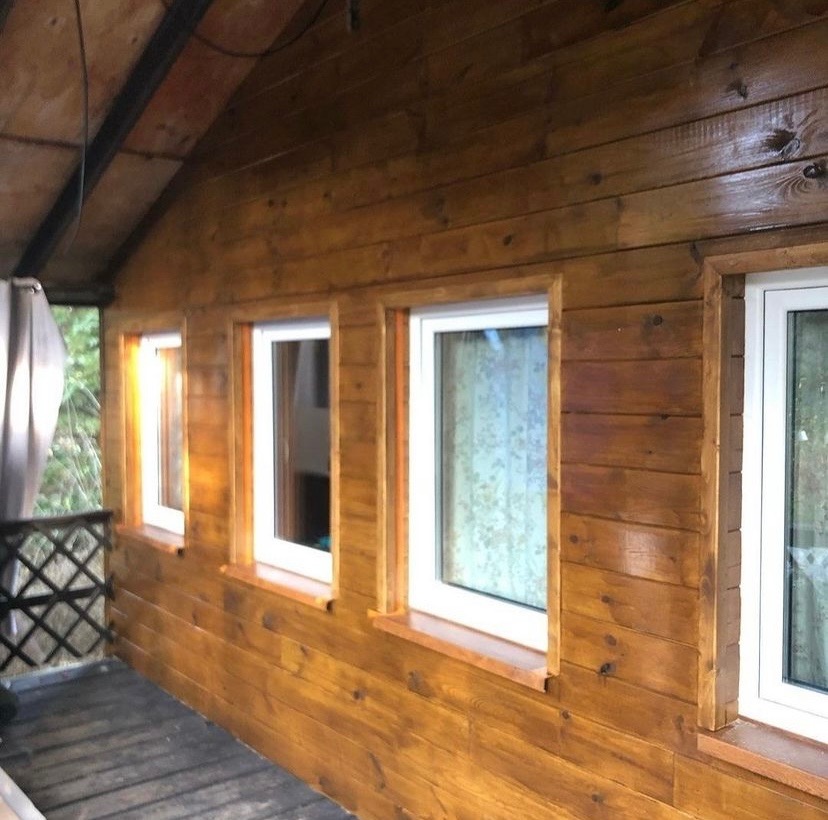 Wnętrze drewnianego domu z trzema oknami z białymi ramami, widok na zewnątrz z zasłoną i balustradą.