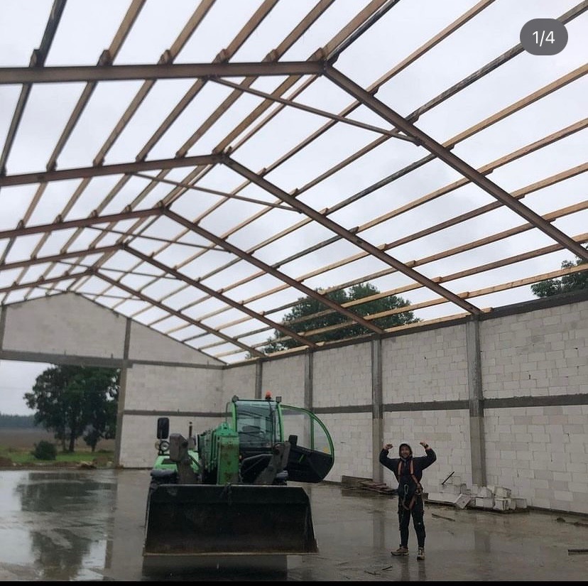 Konstrukcja dachu hali w trakcie budowy, widoczna więźba dachowa z belek, bloczki betonowe ścian i pracownik w uprzęży oraz zielona ładowarka teleskopowa.