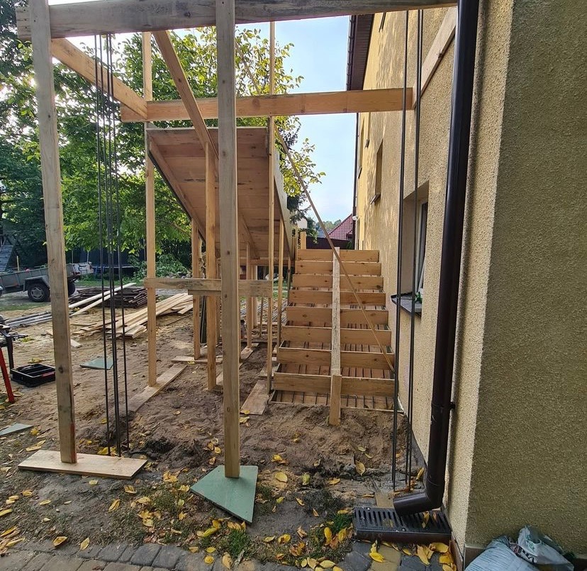 Surowy szkielet drewniany dobudówki z betonowymi schodami, widoczne zbrojenie i rynna przy elewacji budynku.