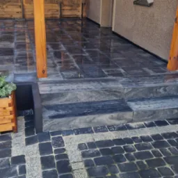 Elegancki taras z ciemnej kostki brukowej i kamiennymi schodami, drewniane donice z białymi kwiatami i dekoracyjne oświetlenie.