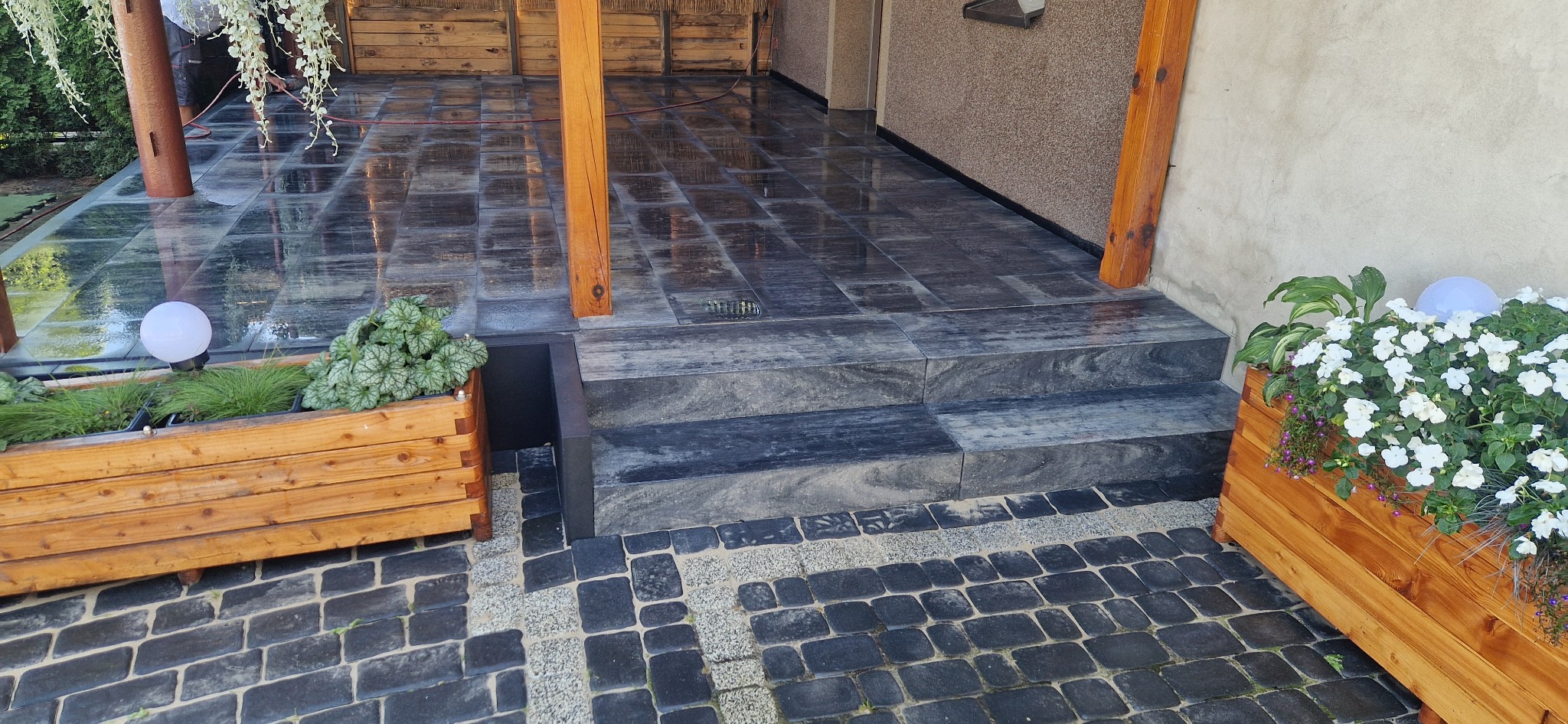 Elegancki taras z ciemnej kostki brukowej i kamiennymi schodami, drewniane donice z białymi kwiatami i dekoracyjne oświetlenie.