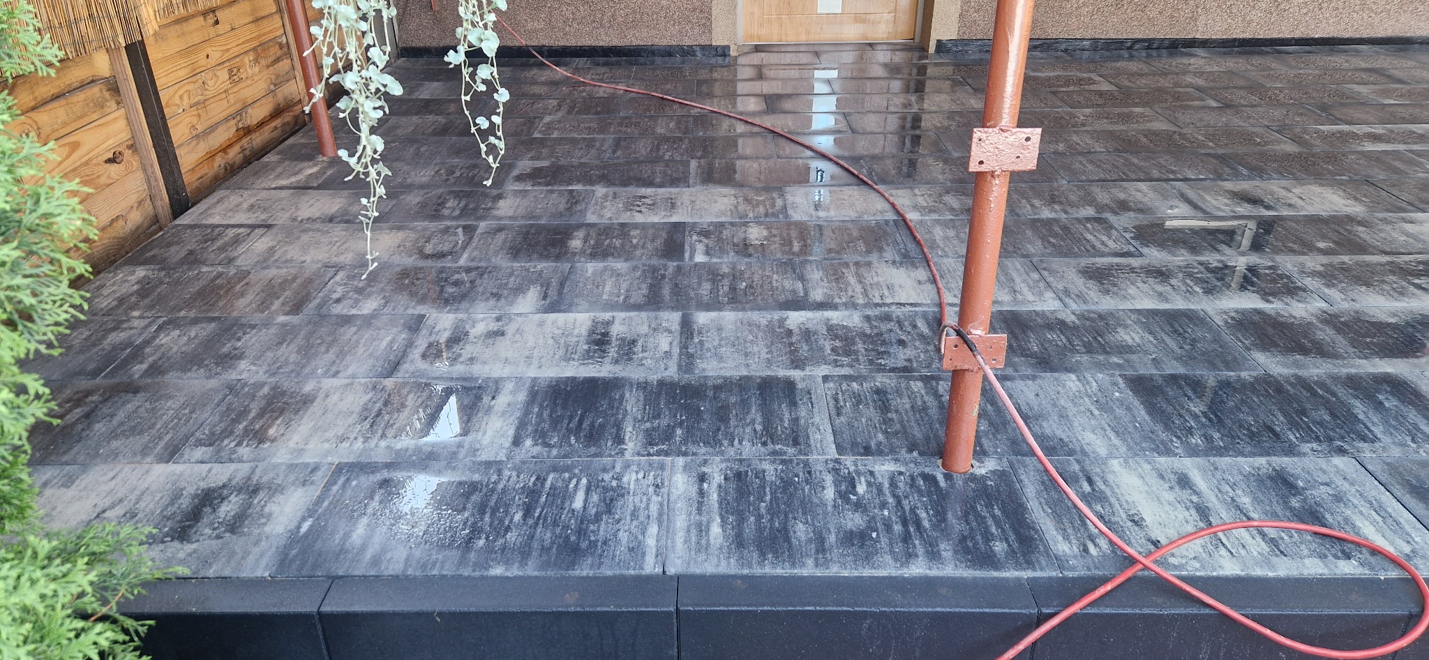 Wyłożony kostką taras w Boronowie, z widocznymi śladami wody i czerwoną rurą zabezpieczającą, z roślinami po bokach.