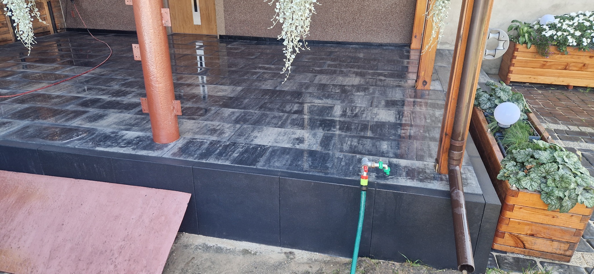 Wykończony taras z kamienną posadzką w odcieniach szarości, zdobiony wiszącymi roślinami, drewnianymi słupami i donicami z kwiatami, z kranem i wężem ogrodowym przy podstawie.