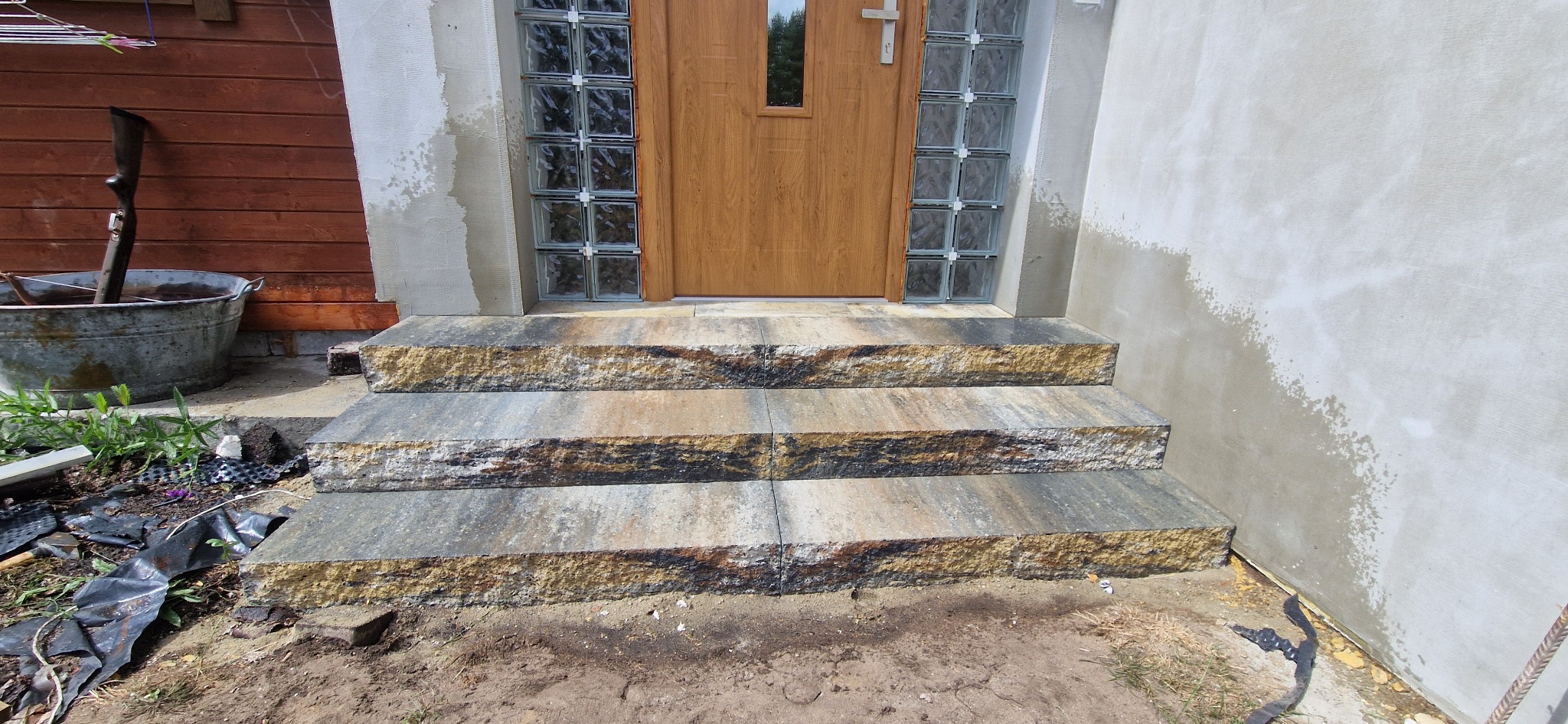 Trzy stopnie schodów z kamiennych płyt prowadzące do drzwi wejściowych, z widocznymi śladami świeżo wykonanych prac murarskich na ścianie i niedokończonym terenem wokół.