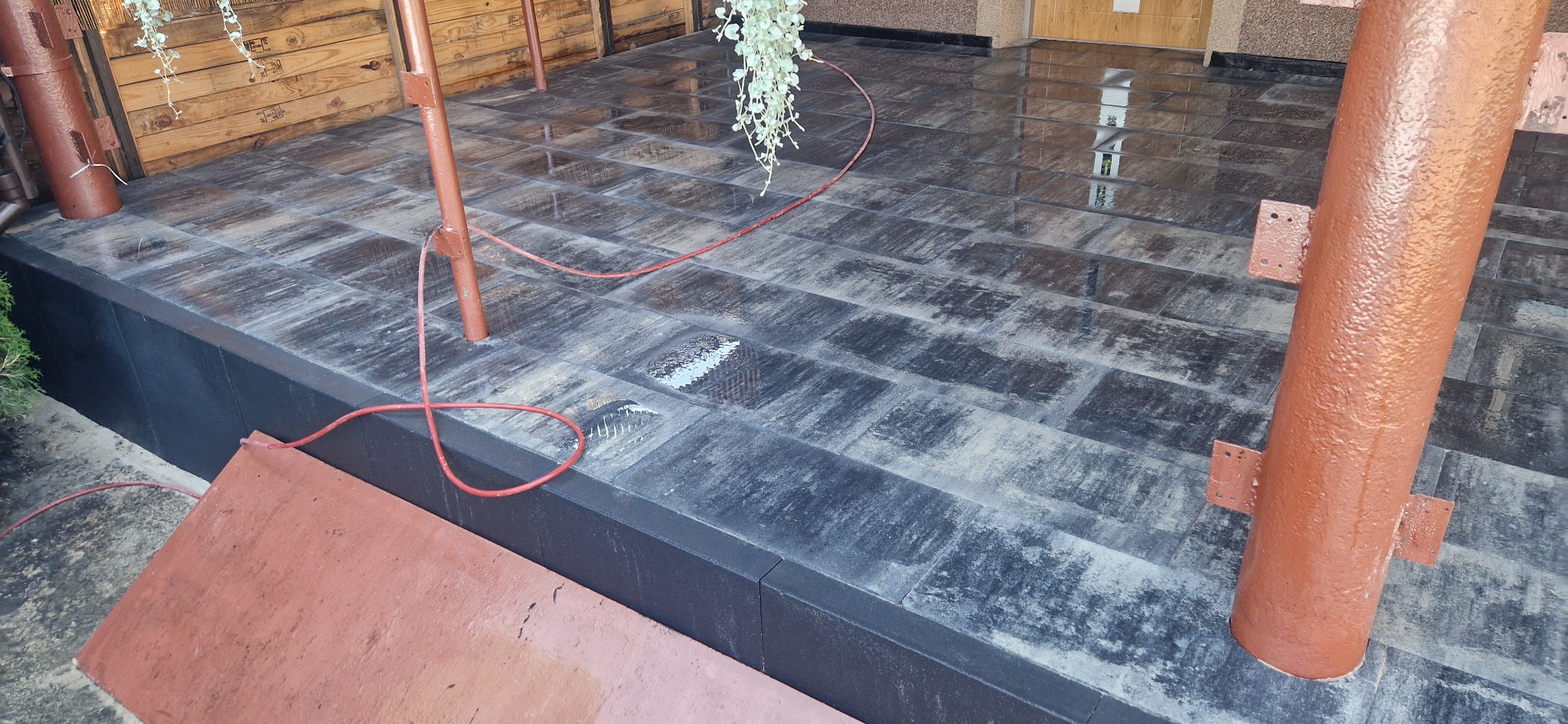 Nowoczesny taras kamienny z ciemnoszarych płyt, częściowo mokry, z drewnianymi elementami konstrukcyjnymi i dekoracyjnymi roślinami wiszącymi.