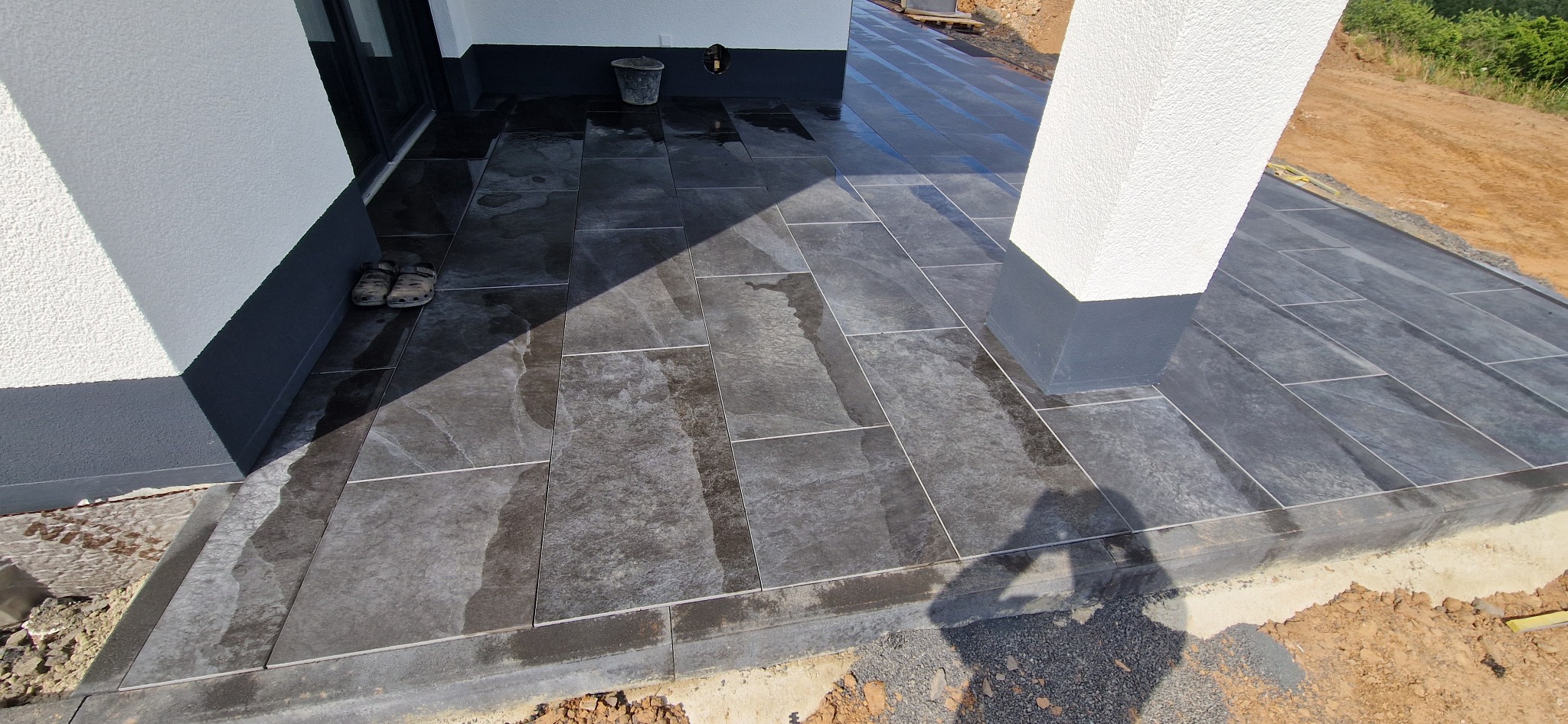 Nowoczesny taras z ciemnoszarych, prostokątnych płyt kamiennych, widok z boku domu z widocznymi butami roboczymi i shadow osoby robiącej zdjęcie.