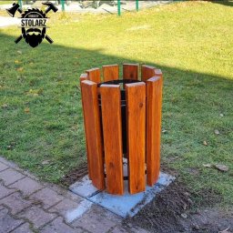 Brodaty stolarz Michał Pajek - Drewniana obudowa kosza na śmieci, wykonana z pionowych desek o ciepłym, brązowym odcieniu, umieszczona na betonowej podstawie na trawniku, z logo firmy w lewym górnym rogu.