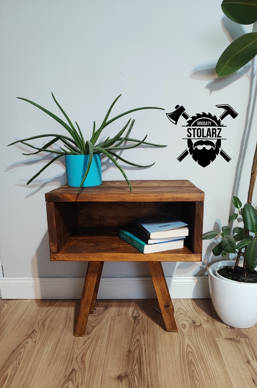 Drewniana szafka nocna z trzema nogami, w stylu retro, z książkami w półce, obok doniczka z aloesem i roślina w białej doniczce, na ścianie naklejka z logo firmy.