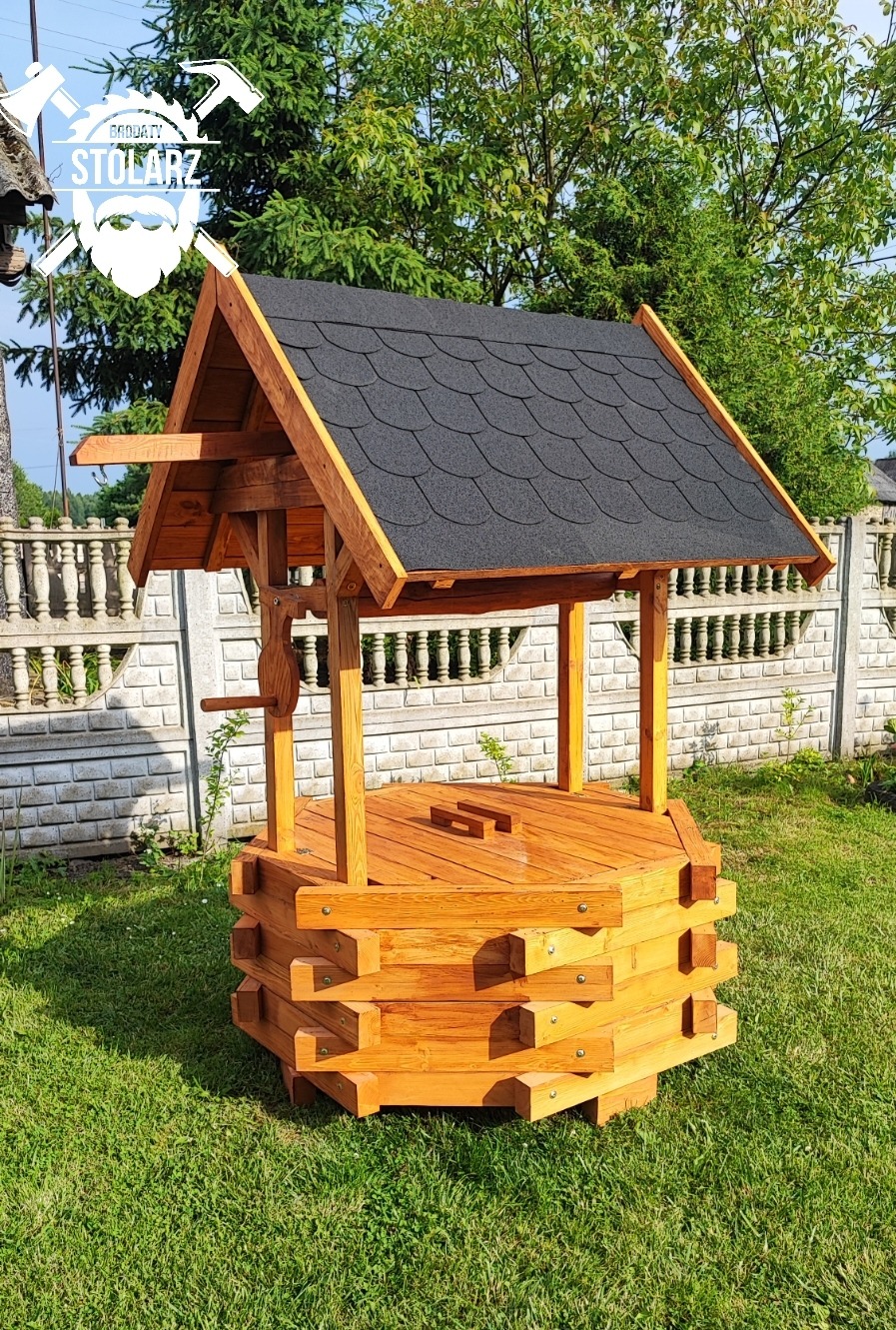 Drewniana studnia ogrodowa z czarnym dachem bitumicznym, umieszczona na trawniku przed kamiennym ogrodzeniem. Widoczny logotyp firmy 'Produkty Stolarz' w lewym górnym rogu zdjęcia.