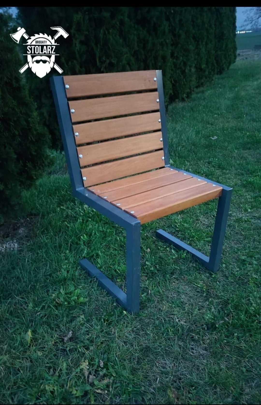 Nowoczesne krzesło ogrodowe z drewnianym siedziskiem i metalową ramą, umieszczone na trawniku przed żywopłotem, z widocznym logo firmy w lewym górnym rogu.