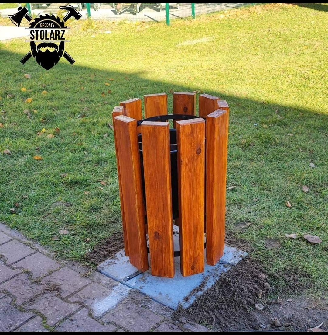 Drewniana obudowa kosza na śmieci, wykonana z pionowych desek o ciepłym, brązowym odcieniu, umieszczona na betonowej podstawie na trawniku, z logo firmy w lewym górnym rogu.