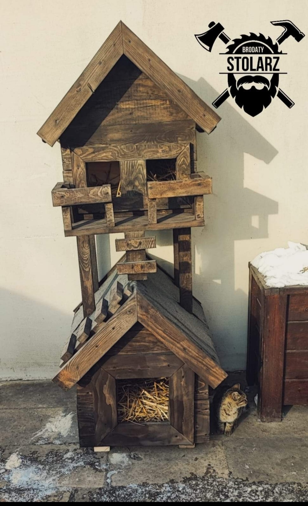Dwupoziomowy, drewniany domek dla zwierząt z sianem w środku, obok siedzi kot, w tle ściana i logo firmy.