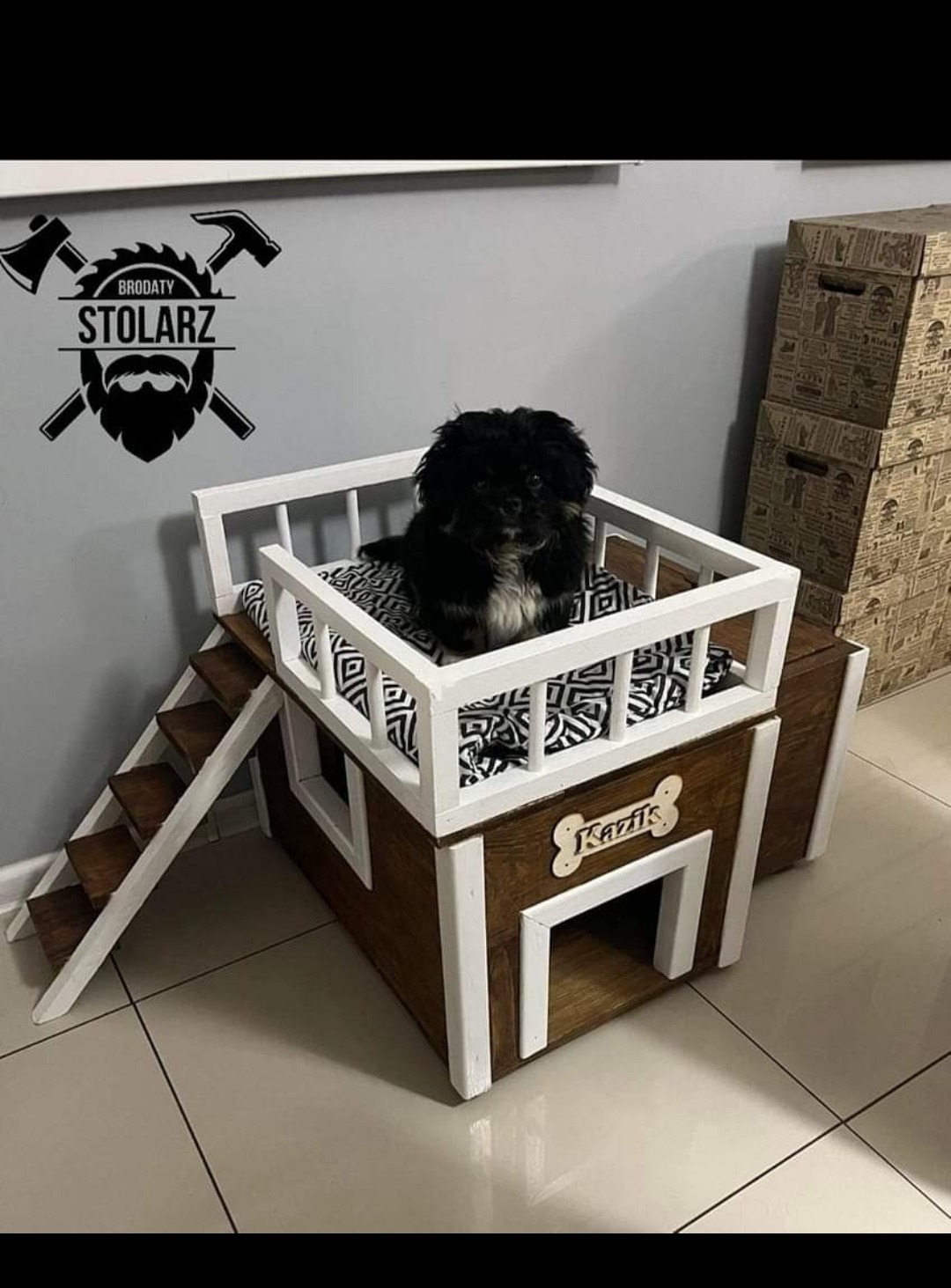 Pies rasy pekińczyk siedzi wewnątrz drewnianego, dwupoziomowego domku dla zwierząt z białymi balustradami i schodkami, obok widoczne logo firmy z narzędziami.
