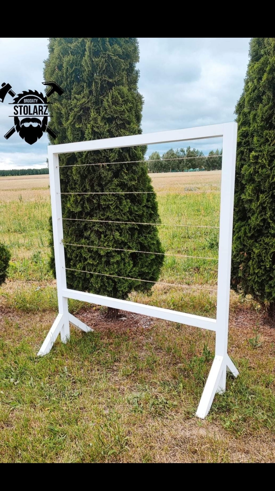 Biała, drewniana konstrukcja z poziomymi sznurkami, ustawiona na trawie przed dwoma wysokimi tujami, z logo firmy w lewym górnym rogu.