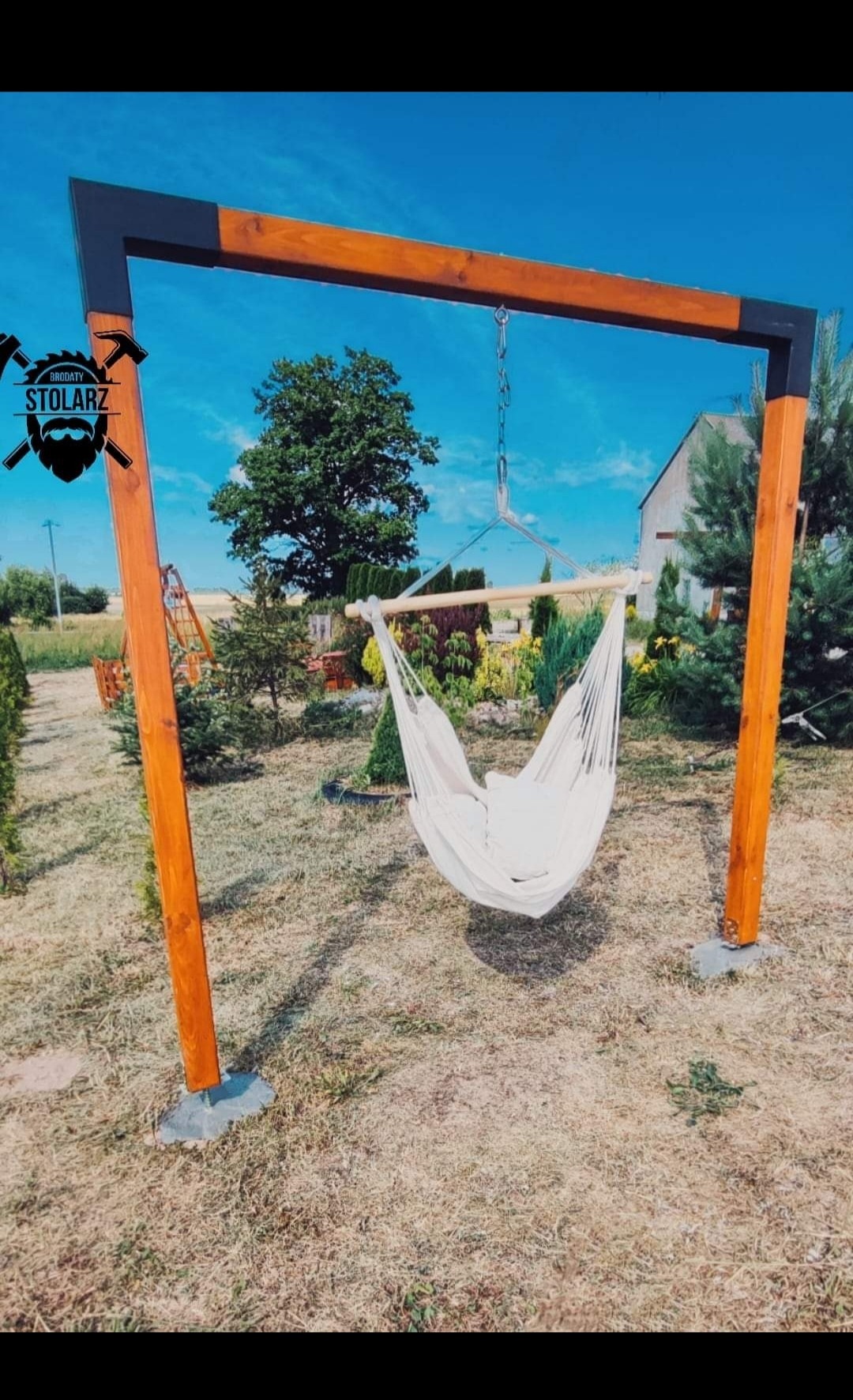 Drewniana konstrukcja huśtawki hamakowej z jasnym hamakiem w ogrodzie, widoczny logo firmy na górze po lewej stronie.