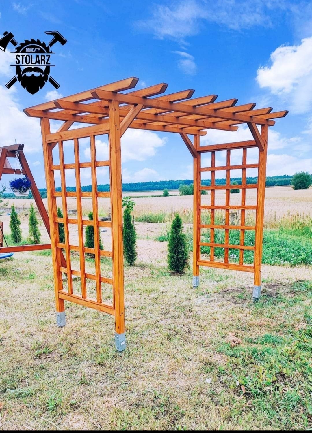 Drewniana pergola z kratownicami, świeżo zaimpregnowana, osadzona na betonowych stopach na trawniku, z logo stolarza w lewym górnym rogu, w tle pole i błękitne niebo.
