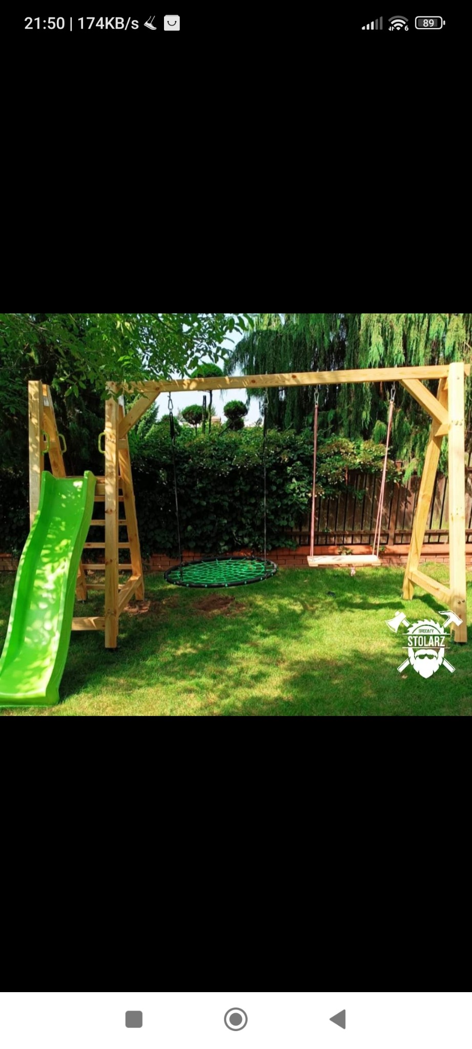 Drewniany plac zabaw z zieloną zjeżdżalnią, okrągłą huśtawką typu 'gniazdo' z zieloną siatką i klasyczną huśtawką na tle zielonego trawnika i żywopłotu, w prawym dolnym rogu widoczne logo firmy.