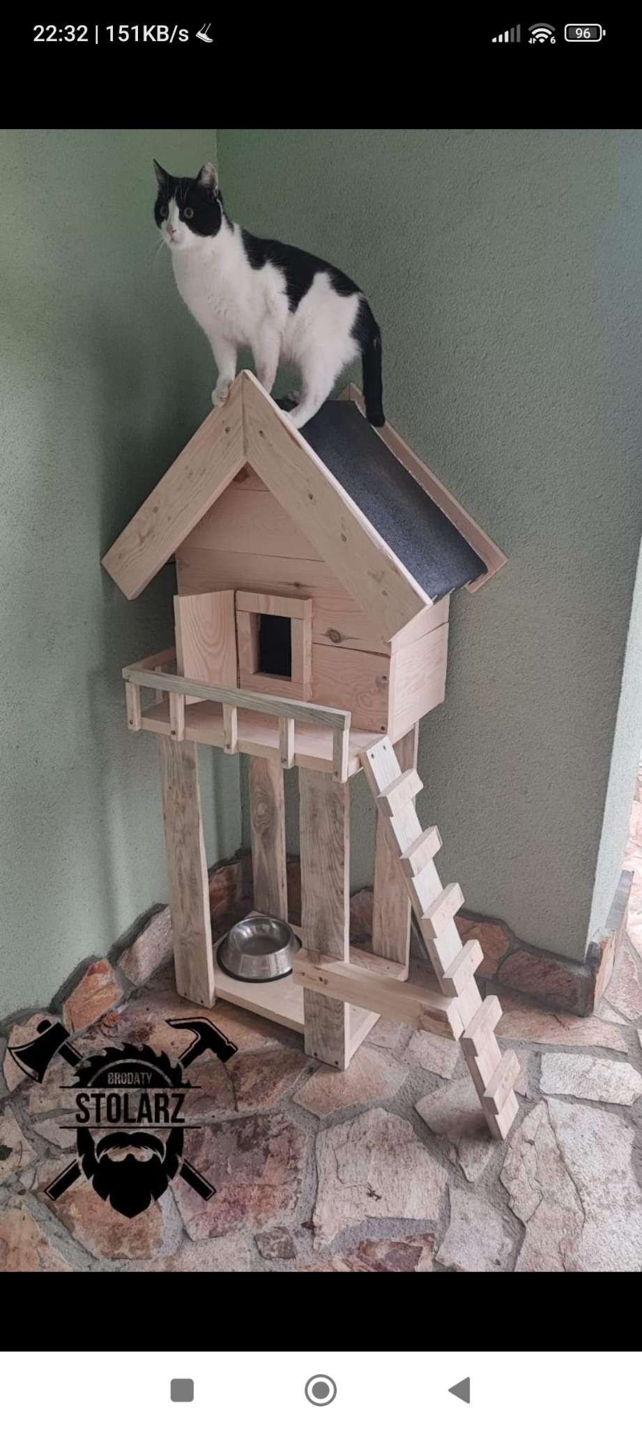 Drewniany domek dla kota z drabinką i miską, na którym stoi czarno-biały kot. W tle widoczne logo firmy.