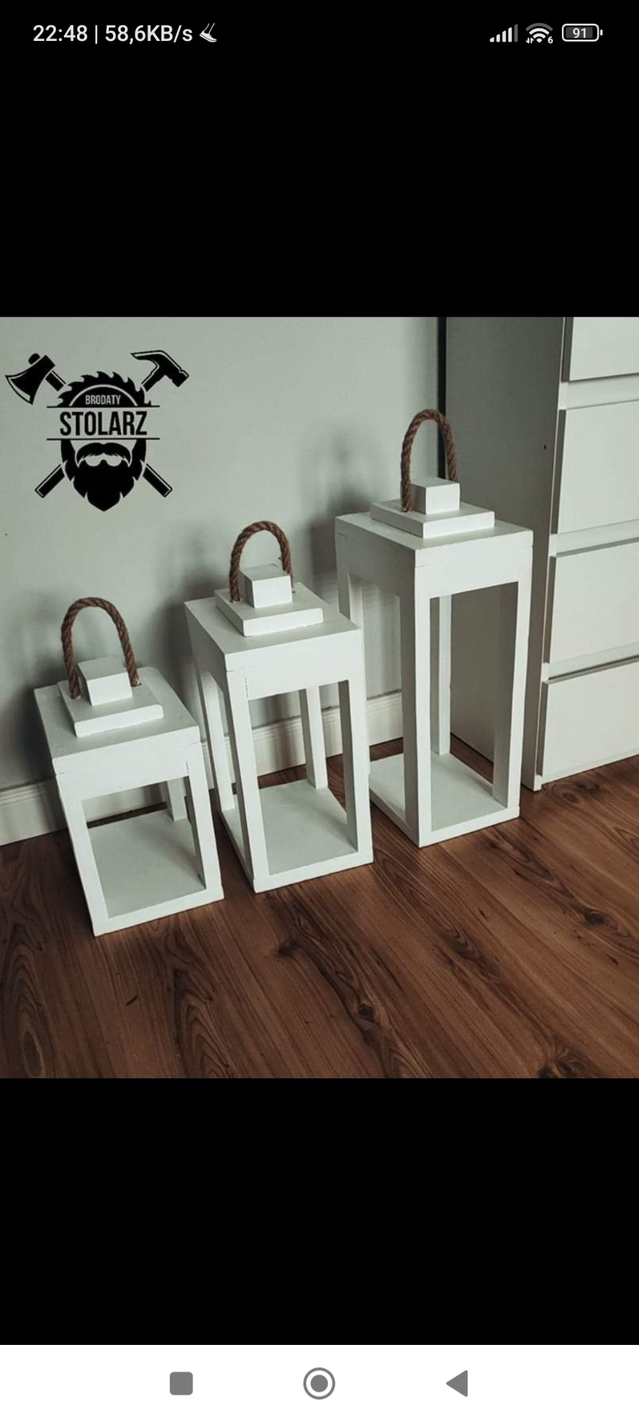 Trzy białe, drewniane lampiony różnej wielkości z uchwytami z grubego sznura, ustawione na podłodze z paneli, na ścianie naklejka z logo firmy z brodatym mężczyzną i narzędziami.