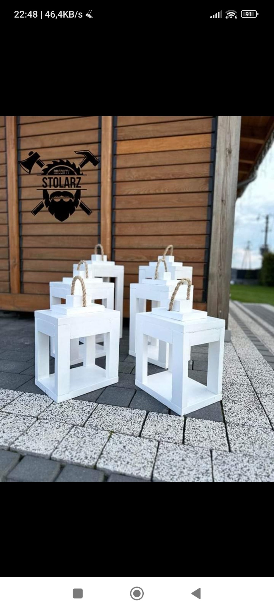 Cztery białe, drewniane lampiony ogrodowe z jutowymi uchwytami, ustawione na kostce brukowej, w tle drewniana ściana z logo firmy stolarskiej.