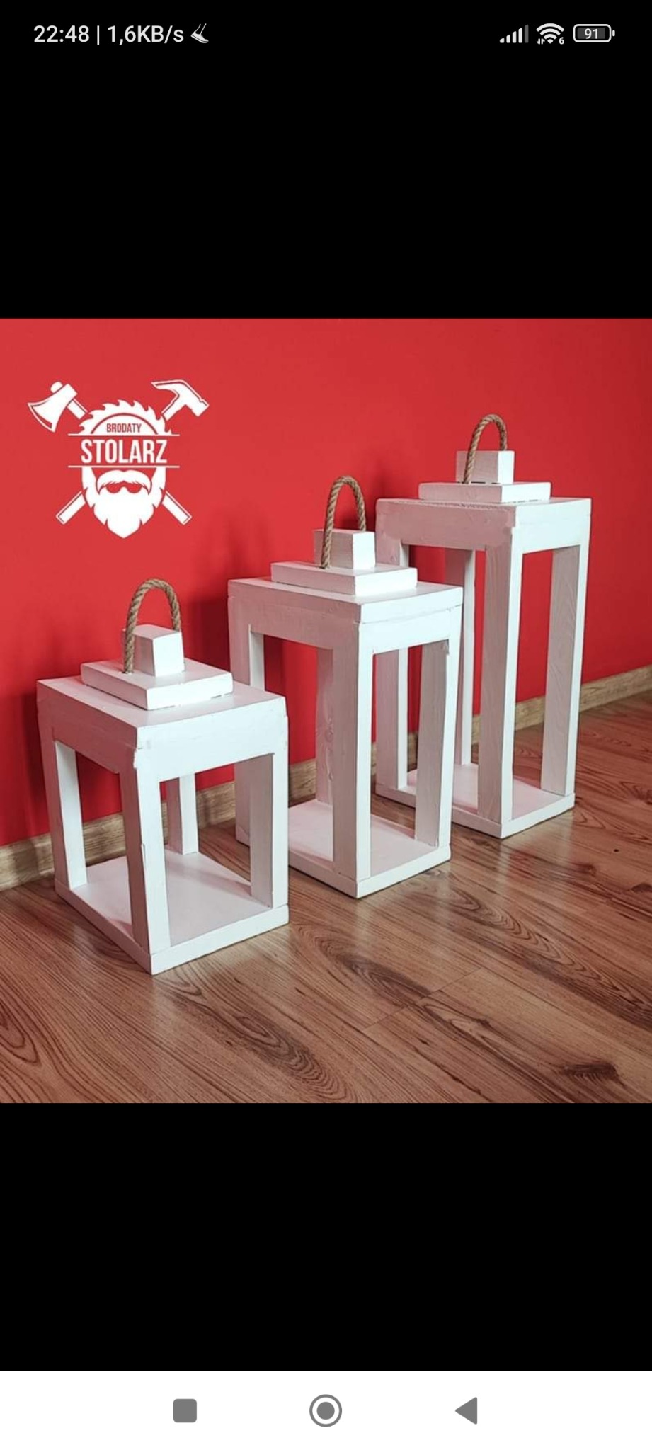 Trzy białe, drewniane lampiony o geometrycznym kształcie z uchwytami z grubego sznura, ustawione na drewnianej podłodze na tle czerwonej ściany, na której widnieje logo firmy z toporami i napisem...