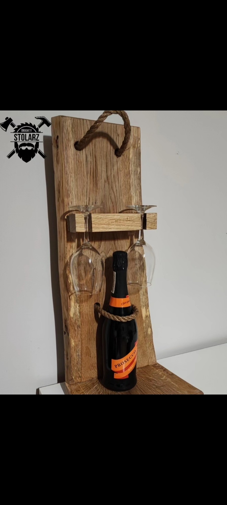 Drewniany stojak na wino i kieliszki z butelką prosecco, zawieszony na ścianie, z widocznym logo firmy w lewym górnym rogu.