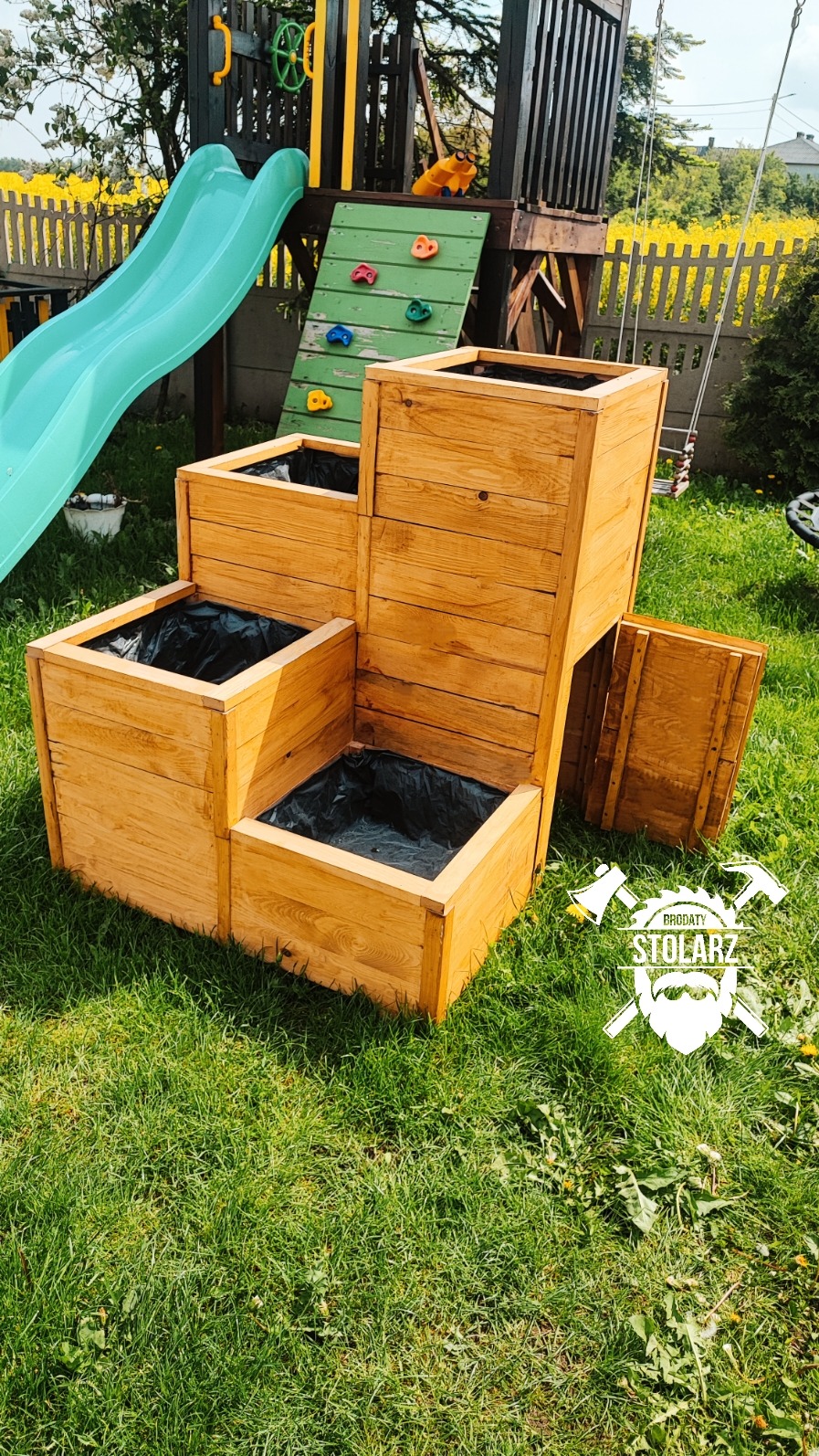 Drewniana, kaskadowa donica ogrodowa z czarnym wkładem, ustawiona na trawie, z placem zabaw w tle i logo stolarza w prawym dolnym rogu.