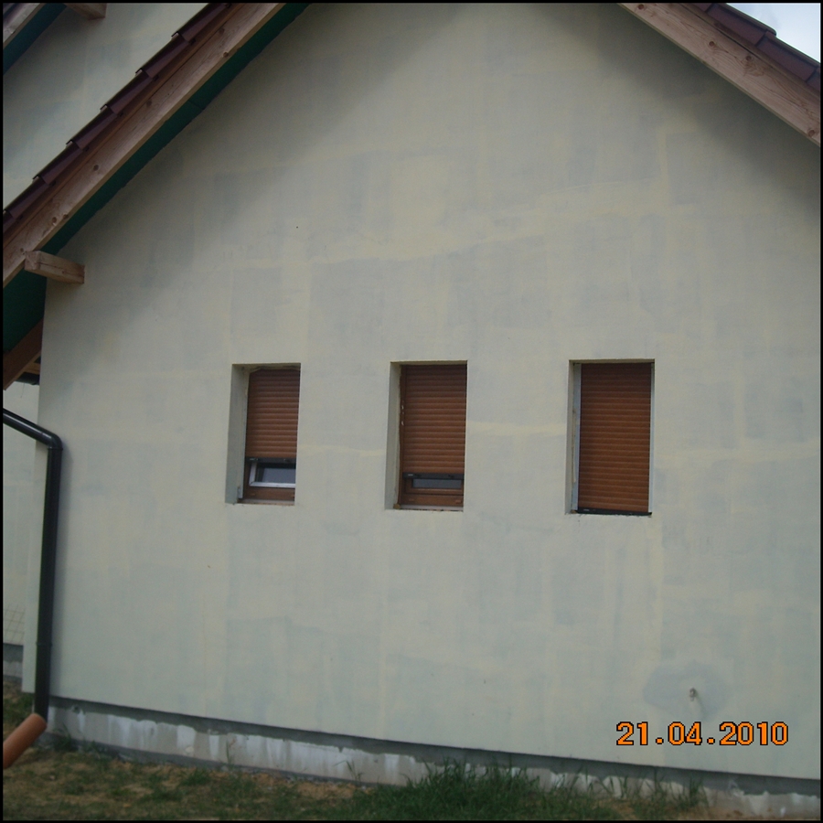 Boczna ściana domu z trzema oknami z roletami zewnętrznymi, świeżo otynkowana i przygotowana do ocieplenia, widoczny fragment dachu i rynny.