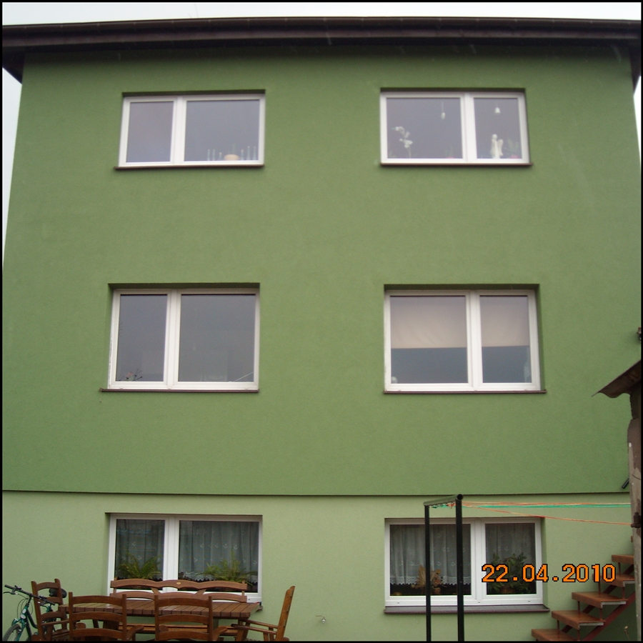 Dwupiętrowy budynek pomalowany na zielono z białymi ramami okiennymi; widoczne meble ogrodowe i schody zewnętrzne.