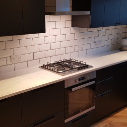 Daniel Demkowski - Nowoczesna kuchnia z czarnymi szafkami, białym blatem imitującym marmur i białymi płytkami typu metro z ciemną fugą. Widoczna płyta gazowa, piekarnik i czajnik elektryczny.