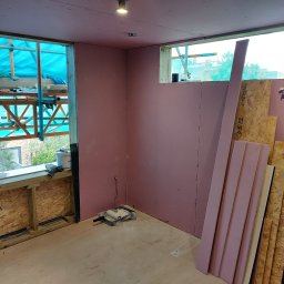 Daniel Demkowski - Wnętrze w trakcie prac wykończeniowych: ściany pokryte różowymi płytami, widoczne profile, okno z rusztowaniem na zewnątrz, narzędzia malarskie na podłodze, przygotowane płyty OSB.