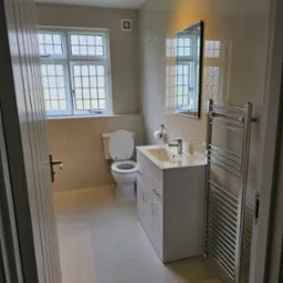 Wykończona łazienka w Bielsku-Białej z beżowymi płytkami, białym zestawem toaletowym, szafką pod umywalką i grzejnikiem drabinkowym, widoczna zza uchylonych drzwi.