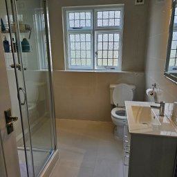 Daniel Demkowski - Jasna łazienka z kabiną prysznicową, umywalką z szafką z szufladami, toaletą i oknem z ozdobnymi szybami, wykończona beżowymi płytkami.