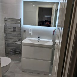 Daniel Demkowski - Nowoczesna łazienka w odcieniach szarości z podświetlanym lustrem nad białą szafką umywalkową, grzejnik drabinkowy po lewej stronie, widok zza uchylonych drzwi.