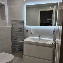 Daniel Demkowski - Wykończona łazienka w odcieniach szarości z podświetlanym lustrem, grzejnikiem drabinkowym i białą szafką pod umywalką, widok na fragment toalety i drzwi.