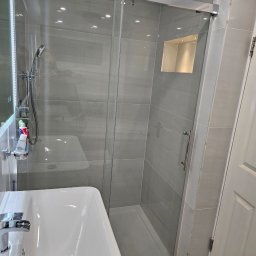 Daniel Demkowski - Nowoczesna łazienka z szarą płytką, kabiną prysznicową ze szklanymi drzwiami przesuwnymi, widoczna umywalka i armatura, oświetlenie halogenowe w suficie i wnęce prysznicowej.