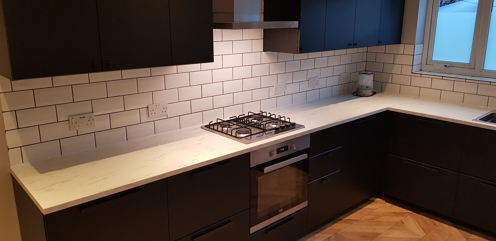 Nowoczesna kuchnia z czarnymi szafkami, białym blatem imitującym marmur i białymi płytkami typu metro z ciemną fugą. Widoczna płyta gazowa, piekarnik i czajnik elektryczny.