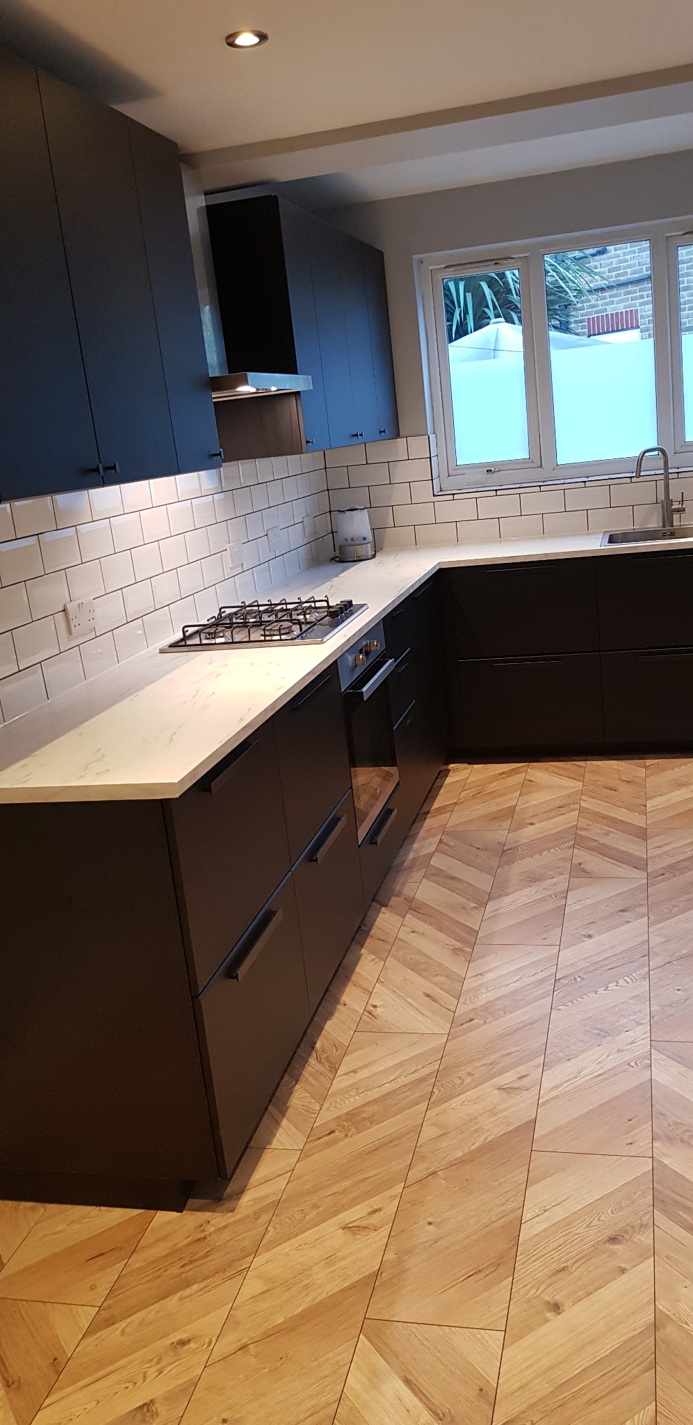 Nowoczesna kuchnia z ciemnymi szafkami, białym blatem, białymi kafelkami na ścianie i parkietem ułożonym w jodełkę.