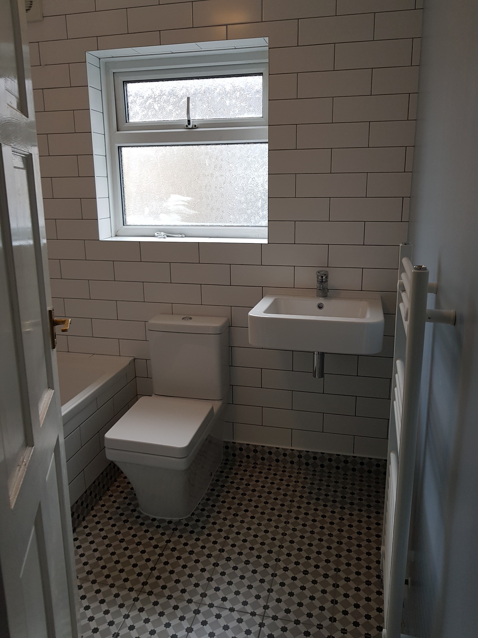 Nowoczesna łazienka po remoncie: białe płytki imitujące cegłę, mozaikowa podłoga, wisząca umywalka, grzejnik drabinkowy i toaleta typu compact.