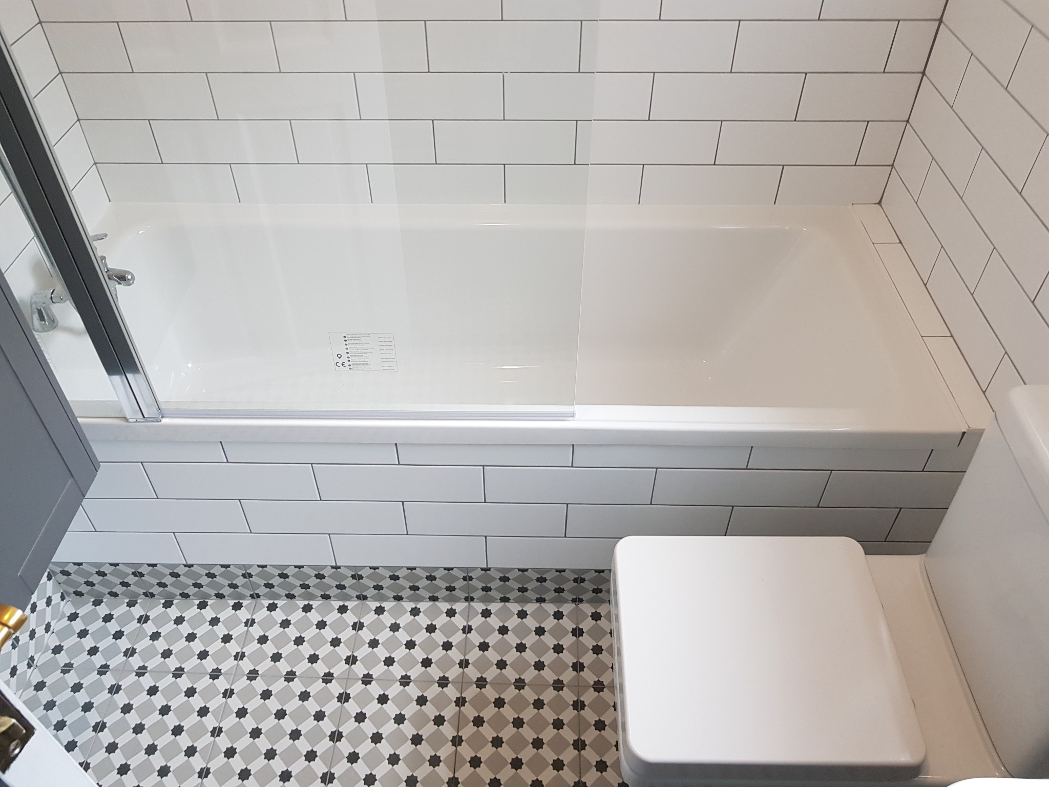 Nowoczesna łazienka z białą wanną z parawanem, białą toaletą i wzorzystą szaro-białą podłogą z płytek.