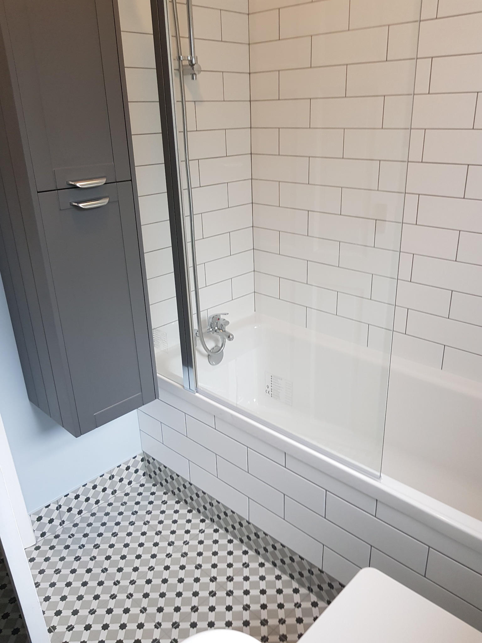 Wanna z zamontowanym szklanym parawanem, obudowana białymi, prostokątnymi płytkami, szara szafka łazienkowa wisząca obok i podłoga wyłożona wzorzystymi płytkami w odcieniach szarości.